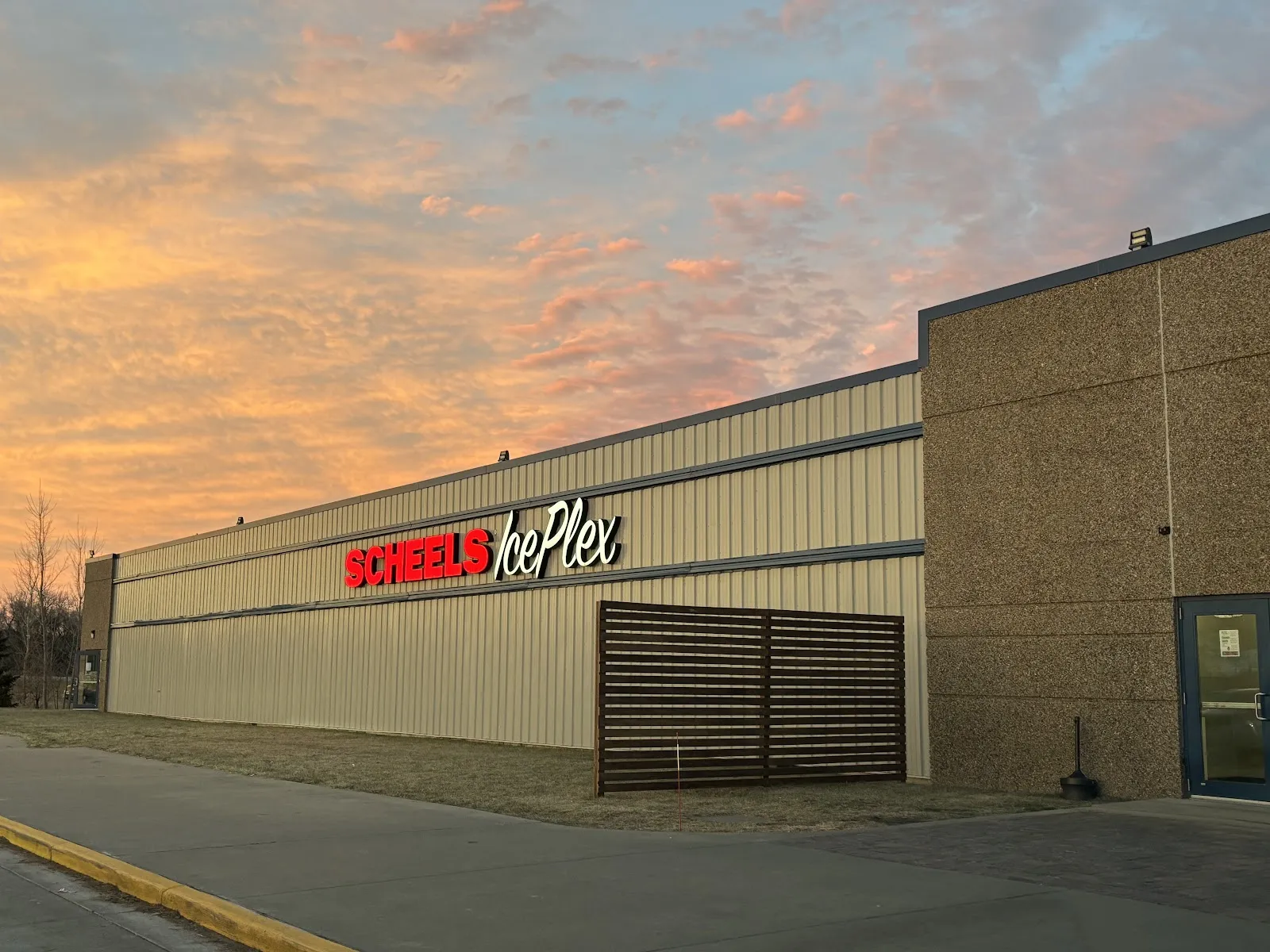 The SCHEELS IcePlex building from the street at sunset, a long tan-brick and metal-clad wall with the SCHEELS IcePlex sign and a slatted wood-look entrance panel under an orange sky. — SCHEELS IcePlex