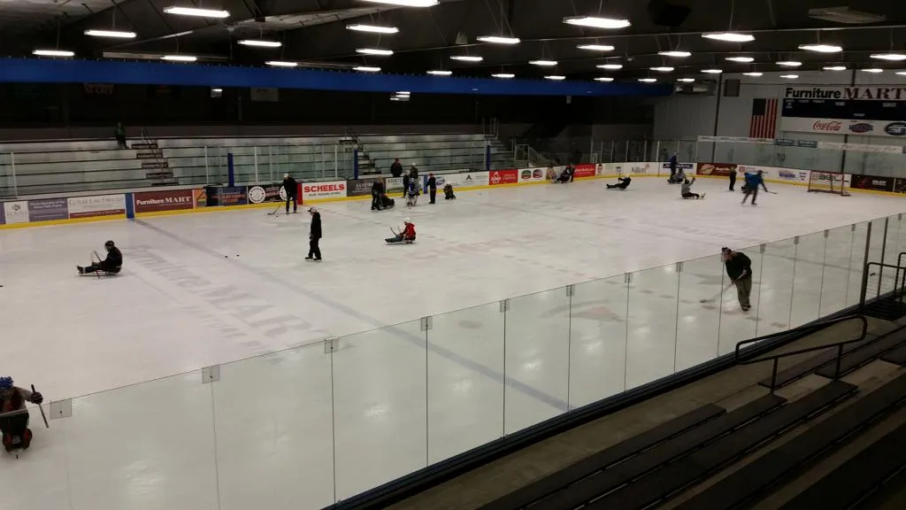 The ice sheet at SCHEELS IcePlex seen from the stands, with glass around the boards, rink-board advertising and a few skaters on the ice — SCHEELS IcePlex