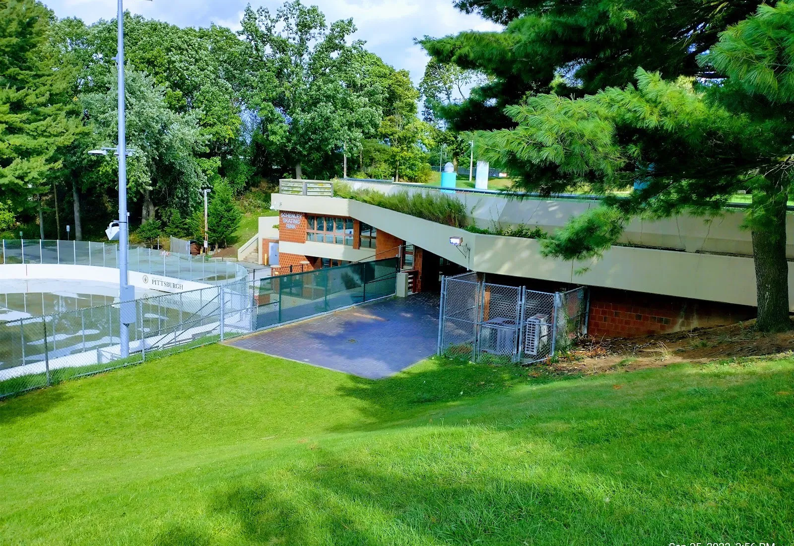 The Schenley Skating Rink building, signed on its brick end and set into the hillside under a green roof, beside the open rink in summer — Schenley Park Skating Rink