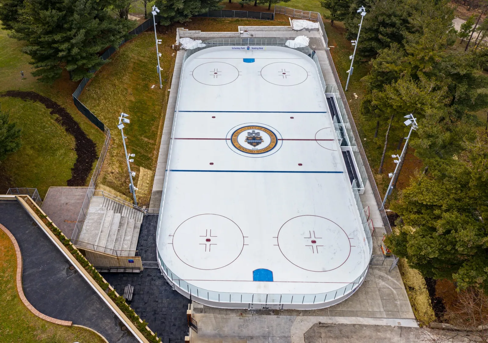 Aerial view of the full outdoor ice sheet at Schenley Park Skating Rink, empty, with hockey lines, the City of Pittsburgh seal painted at center ice, glass dasher boards, and a 'Schenley Park Skating Rink' sign at one end. — Schenley Park Skating Rink
