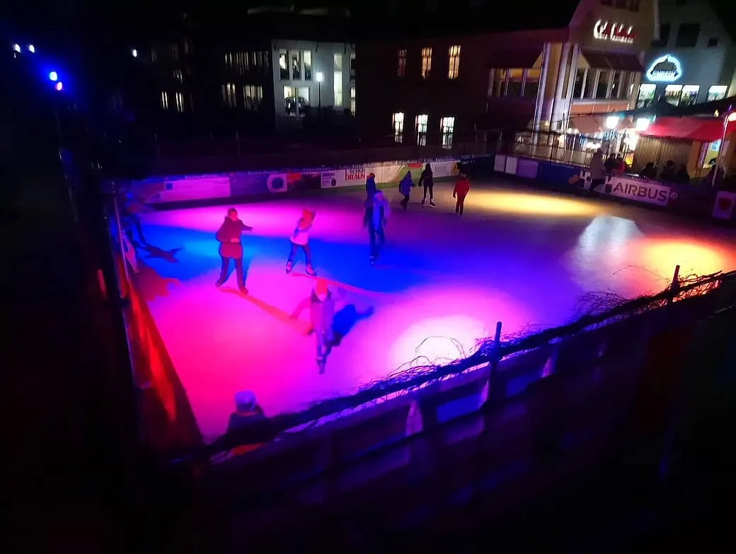 The Plätzle skating rink in Donauwörth at night, lit in pink and blue, with a few skaters and the old town houses around it — Schlittschuhbahn Plätzle Donauwörth