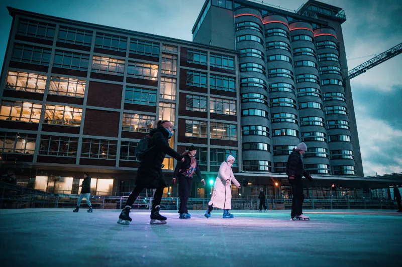 Skaters in a group on the ice at dusk in front of the brick warehouse buildings and round silo tower of the Überseeinsel. — Schlittschuhspaß Überseeinsel