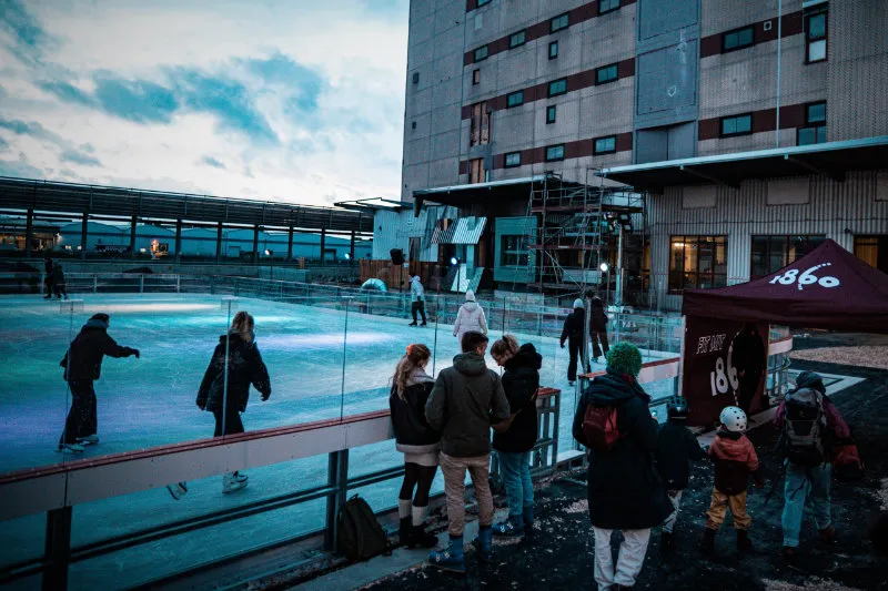 The open-air rink on the Überseeinsel in Bremen at dusk, skaters on the ice and onlookers at the glass boards, with the converted warehouse building behind. — Schlittschuhspaß Überseeinsel