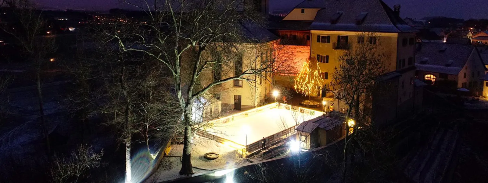 The lit Schloss-Eisbahn in the courtyard of Schloss Grüningen seen from above at night, among the houses of the village — Schloss-Eisbahn Grüningen