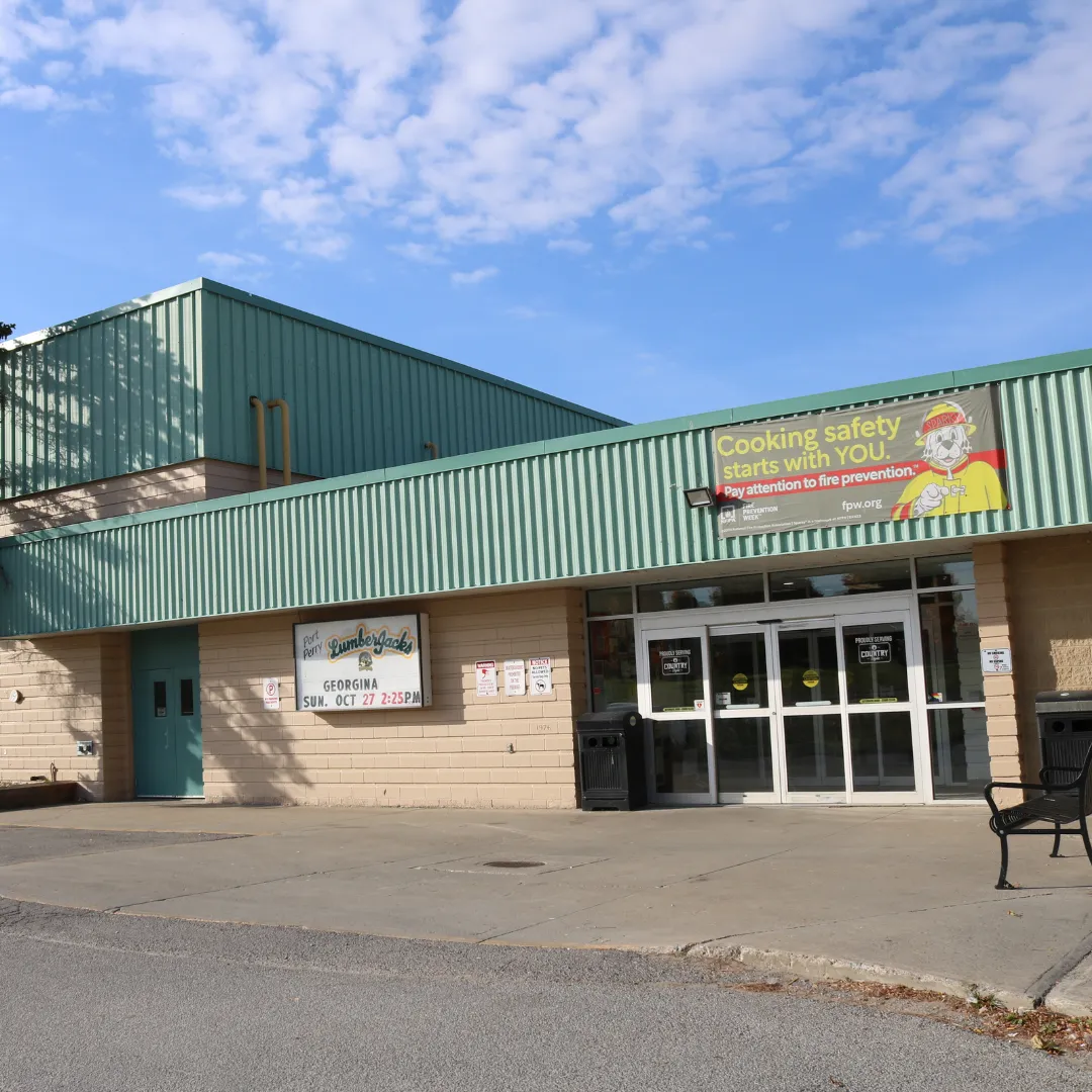 The arena entrance under green metal cladding, with a community sign board beside the glass doors. — Scugog Arena (Port Perry)