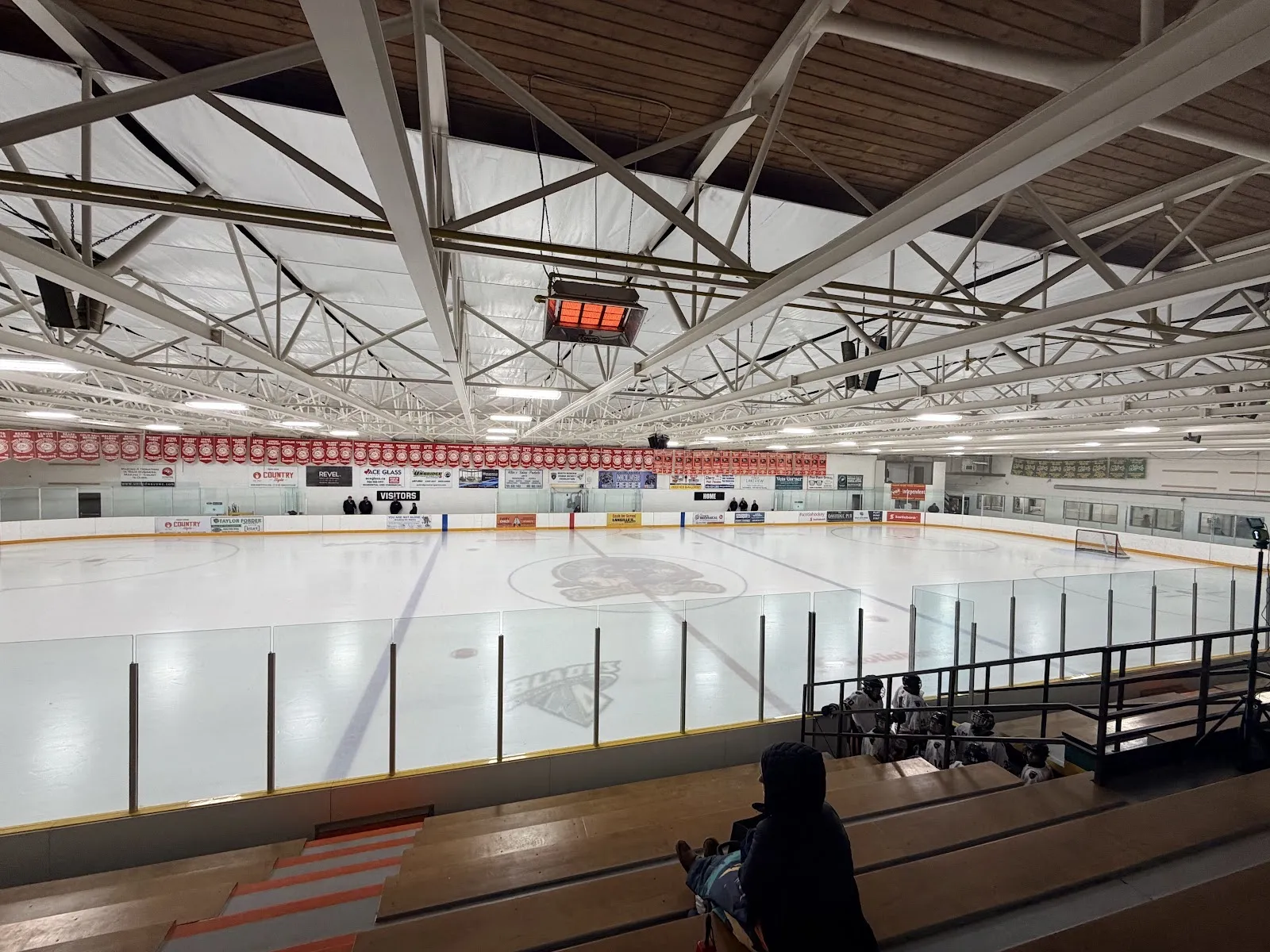 The empty ice with a Blades logo at centre, seen from the wooden bleachers under steel trusses and a row of red banners. — Scugog Arena (Port Perry)
