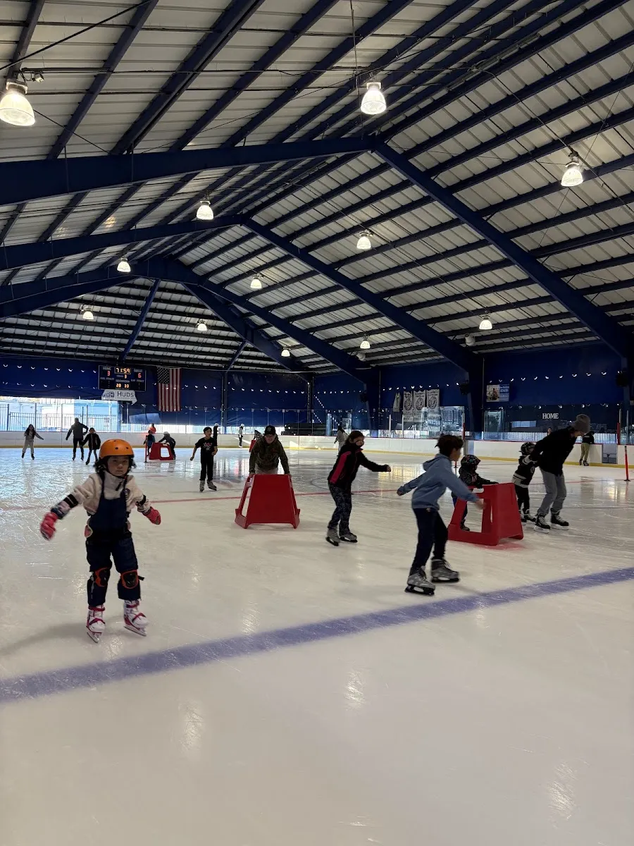 Skaters, some pushing red skating aids, spread across the covered Secaucus rink under a blue steel roof. — Secaucus Ice Rink