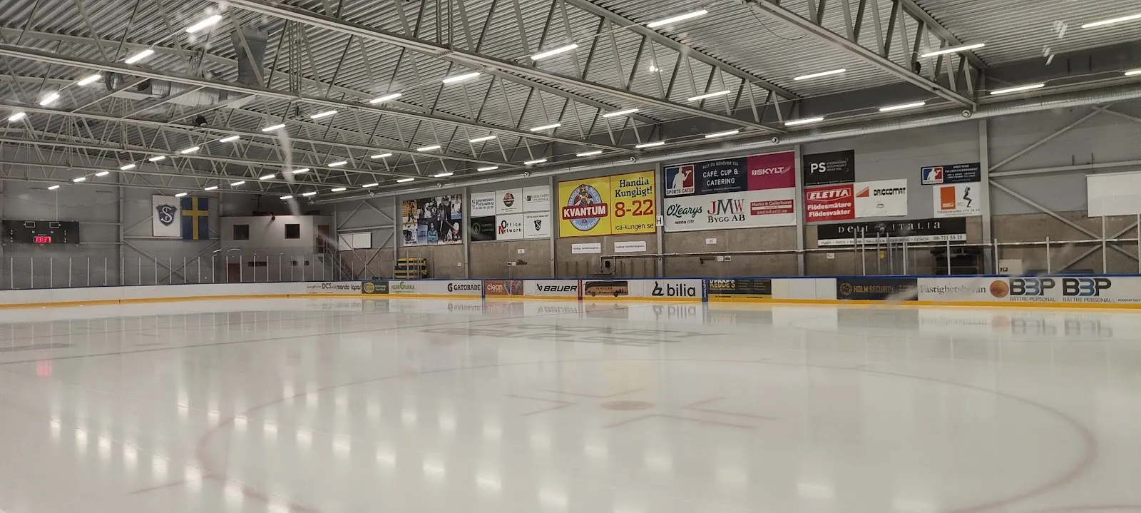 The long empty ice sheet inside Segeltorpshallen, with the boards, the sponsor wall at the far end and the white roof above. — Segeltorpshallen