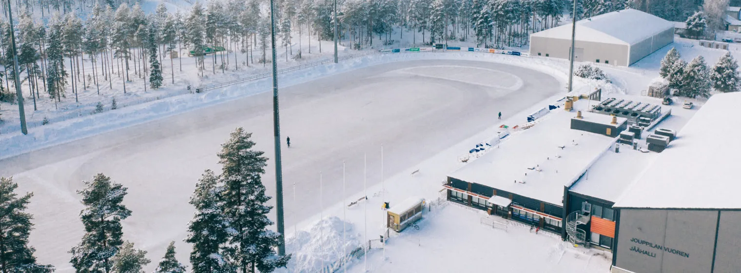 Aerial view of the large artificial-ice track at the Seinäjoki ice sports centre in winter, with snow-covered pines and the arena buildings beside it. — Seinäjoen Iso tekojäärata