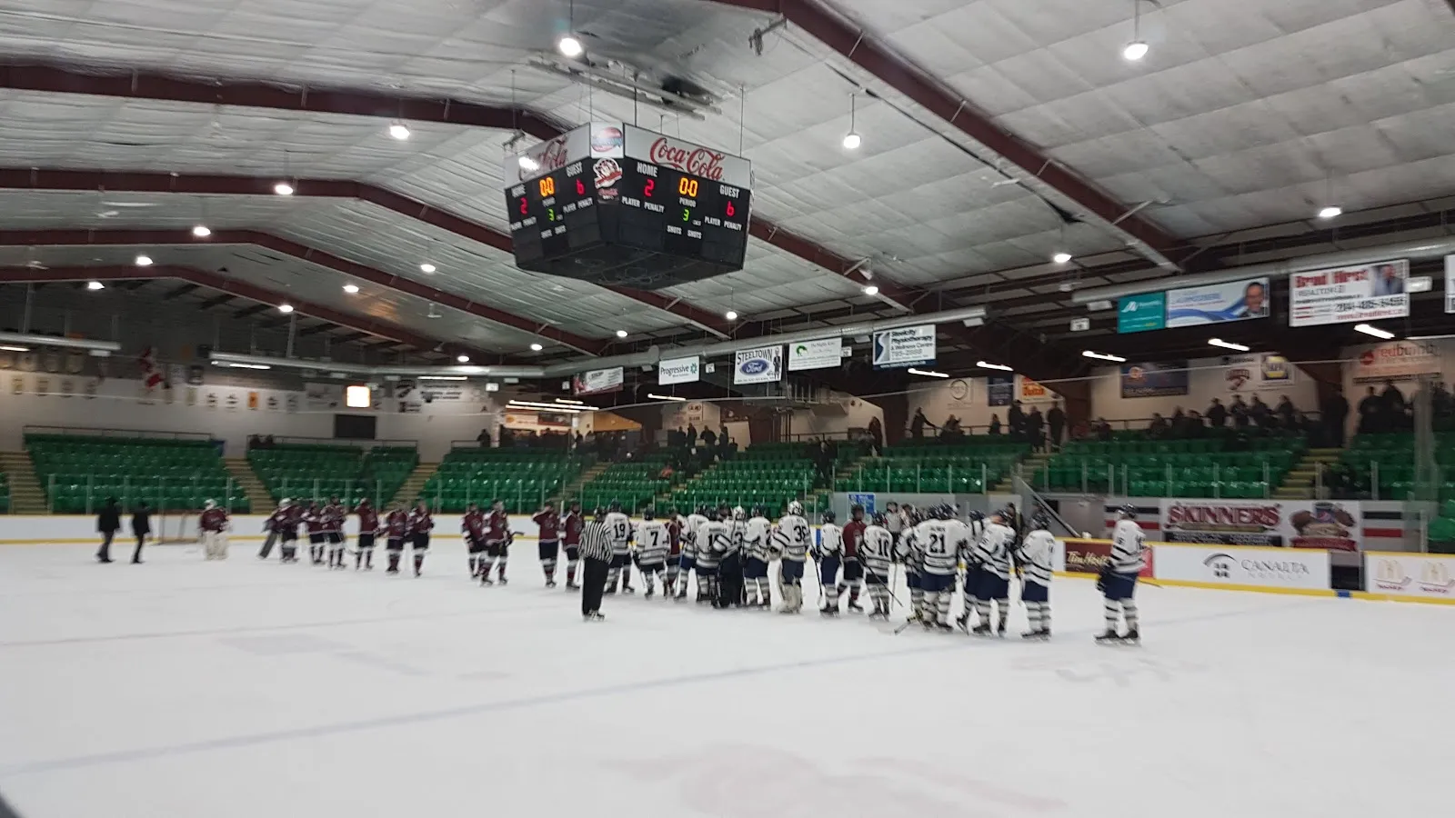 Two teams lined up on the far side of the ice at the Selkirk Recreation Complex, with green seating and a scoreboard overhead. — Selkirk Recreation Complex