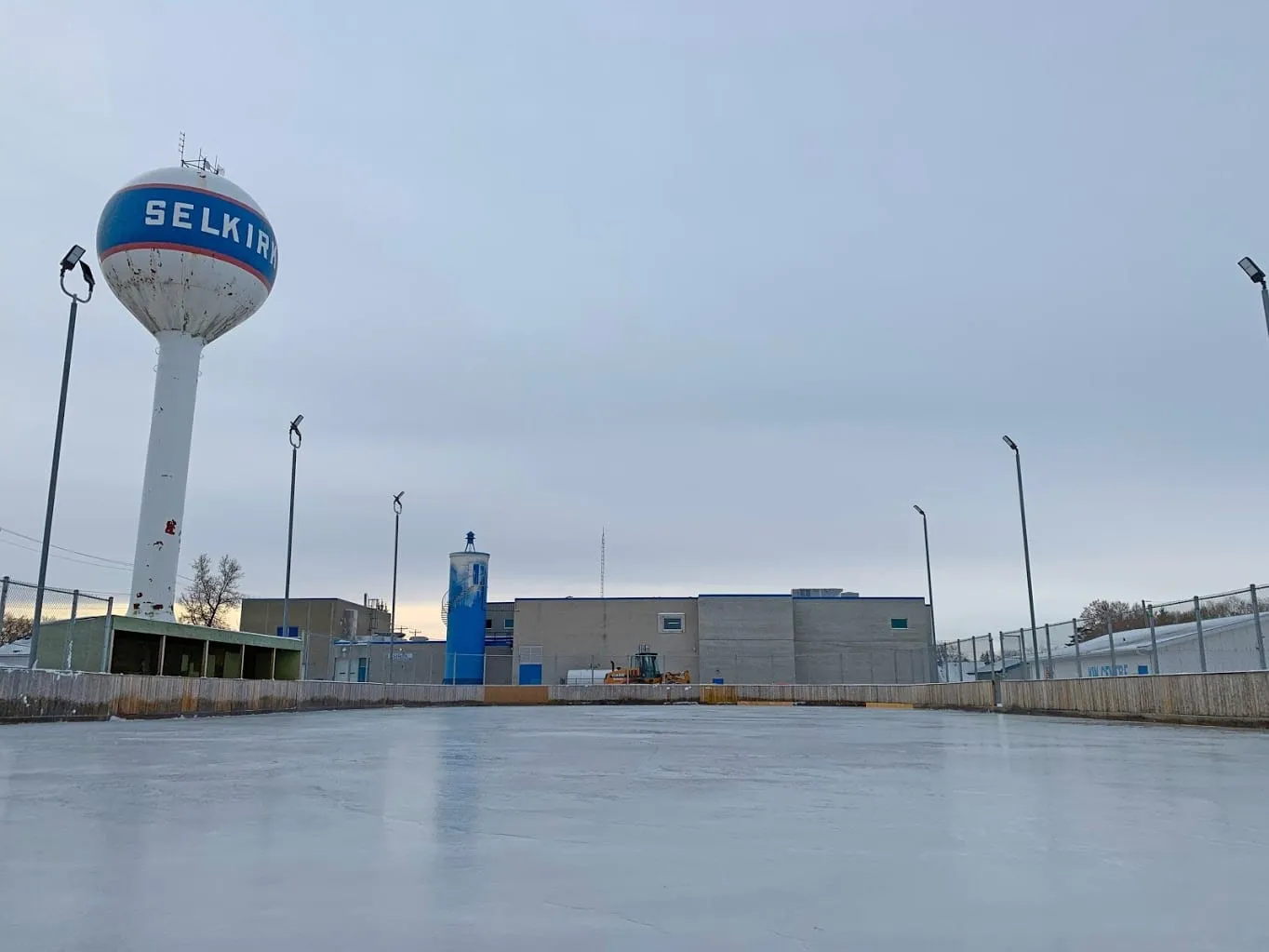 La pista exterior de Water Tower Park en Selkirk bajo un cielo gris de atardecer, con el depósito de agua de la ciudad y los focos más allá de la valla. — Selkirk Water Tower Park Outdoor Rink