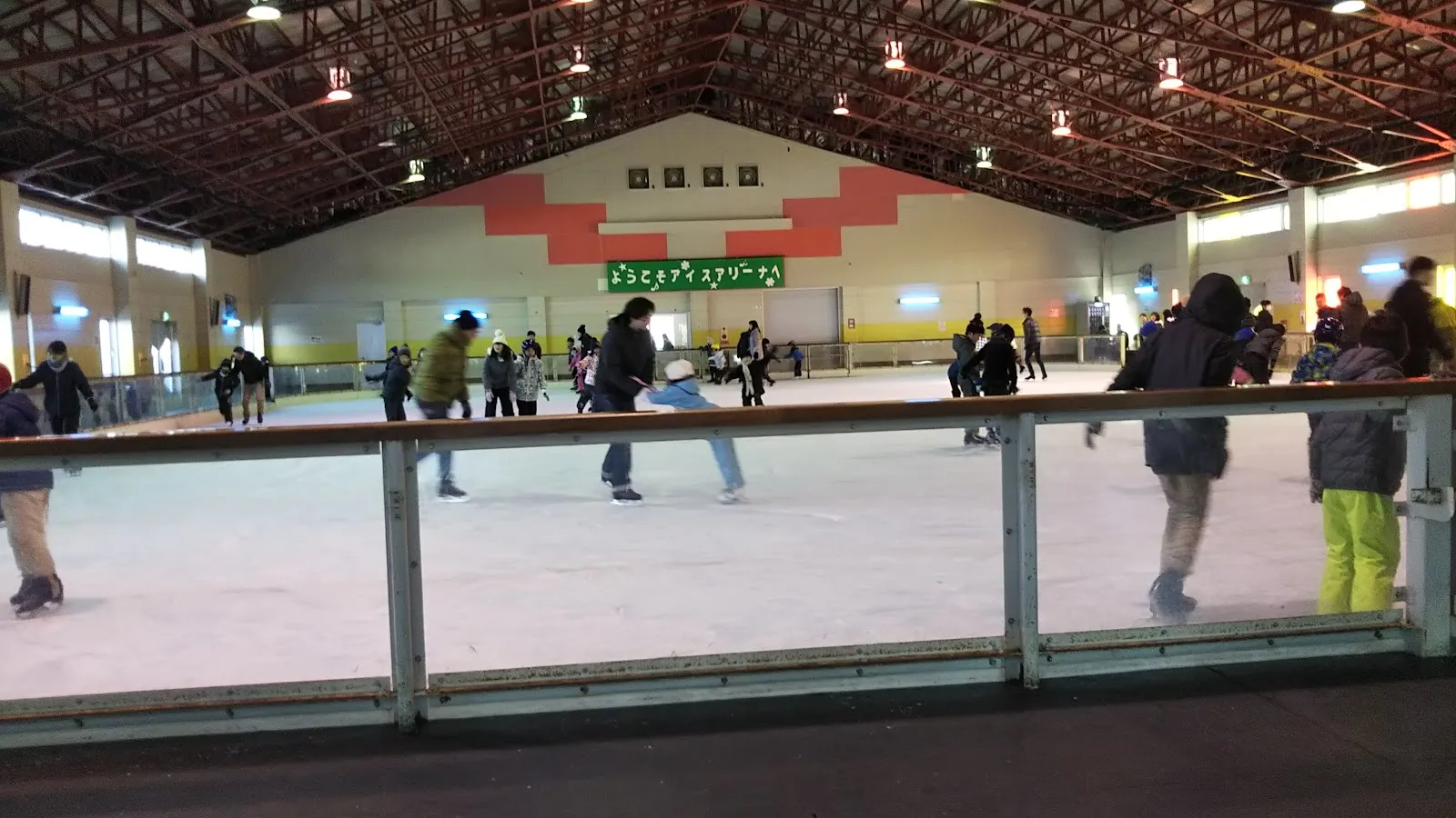 Skaters on the ice of Senmaya Ice Arena seen from behind the boards, under a wooden-roofed hall with a green banner on the far wall. — Senmaya Ice Arena