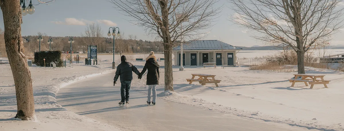 Two skaters seen from behind on the Magog skating trail along the shore in winter, with bare trees, picnic tables and a small building beside the lake. — Sentier glacé de Magog (pointe Merry et pointe Cabana)