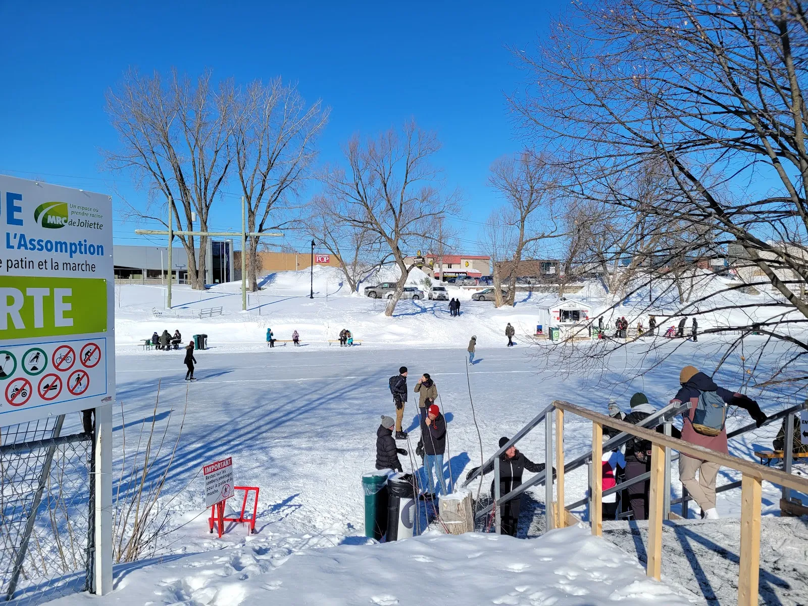The stairs and signboard at the entrance to the river skating trail beside Parc Louis-Querbes in Joliette, with skaters on the ice beyond — Sentier glacé du parc Louis-Querbes