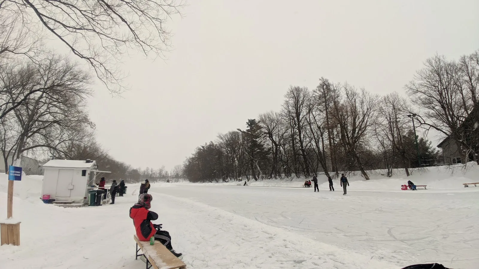 The groomed skating trail on the frozen L'Assomption river in Joliette, with a warming hut, benches and skaters at a distance — Sentier glacé du parc Louis-Querbes