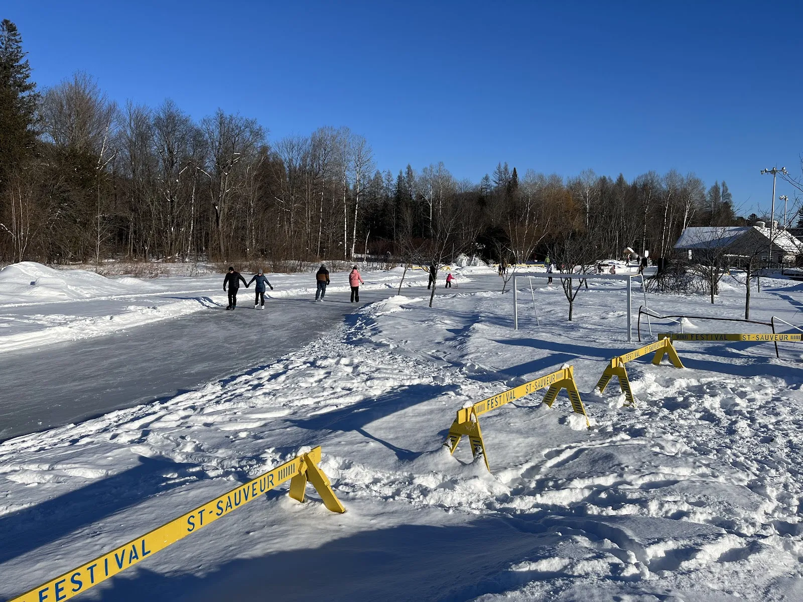 Skaters far along the ice trail at the John-H.-Molson park in Saint-Sauveur on a sunny day, with yellow barriers and the park buildings beyond — Sentiers glacés du parc John-H.-Molson