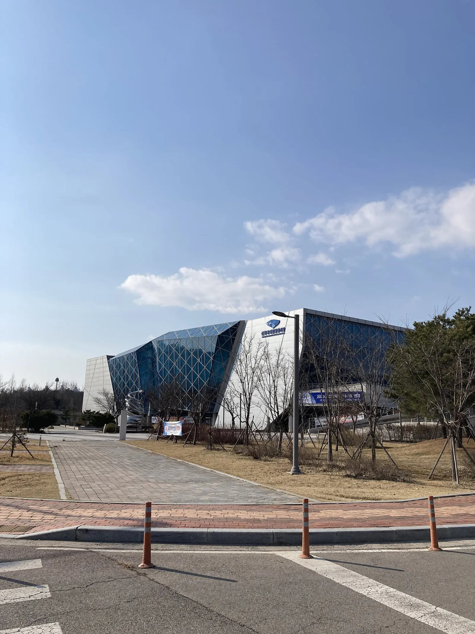 The faceted blue-glass and white building of Seonhak International Ice Rink seen across the plaza from the road, with the entrance banner at its foot. — Seonhak International Ice Rink