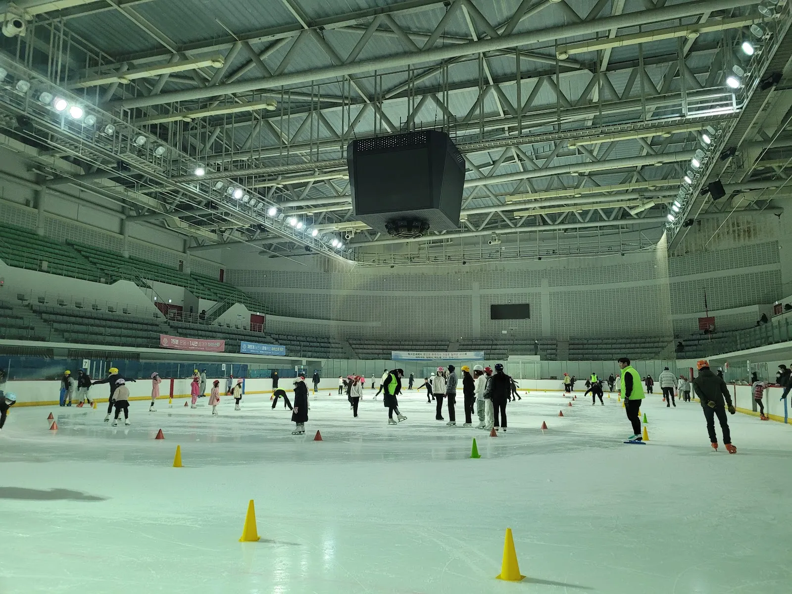 The main rink of Seonhak International Ice Rink from the boards: the steel-trussed hall, the scoreboard and the stands above a public session with small distant skaters among training cones. — Seonhak International Ice Rink