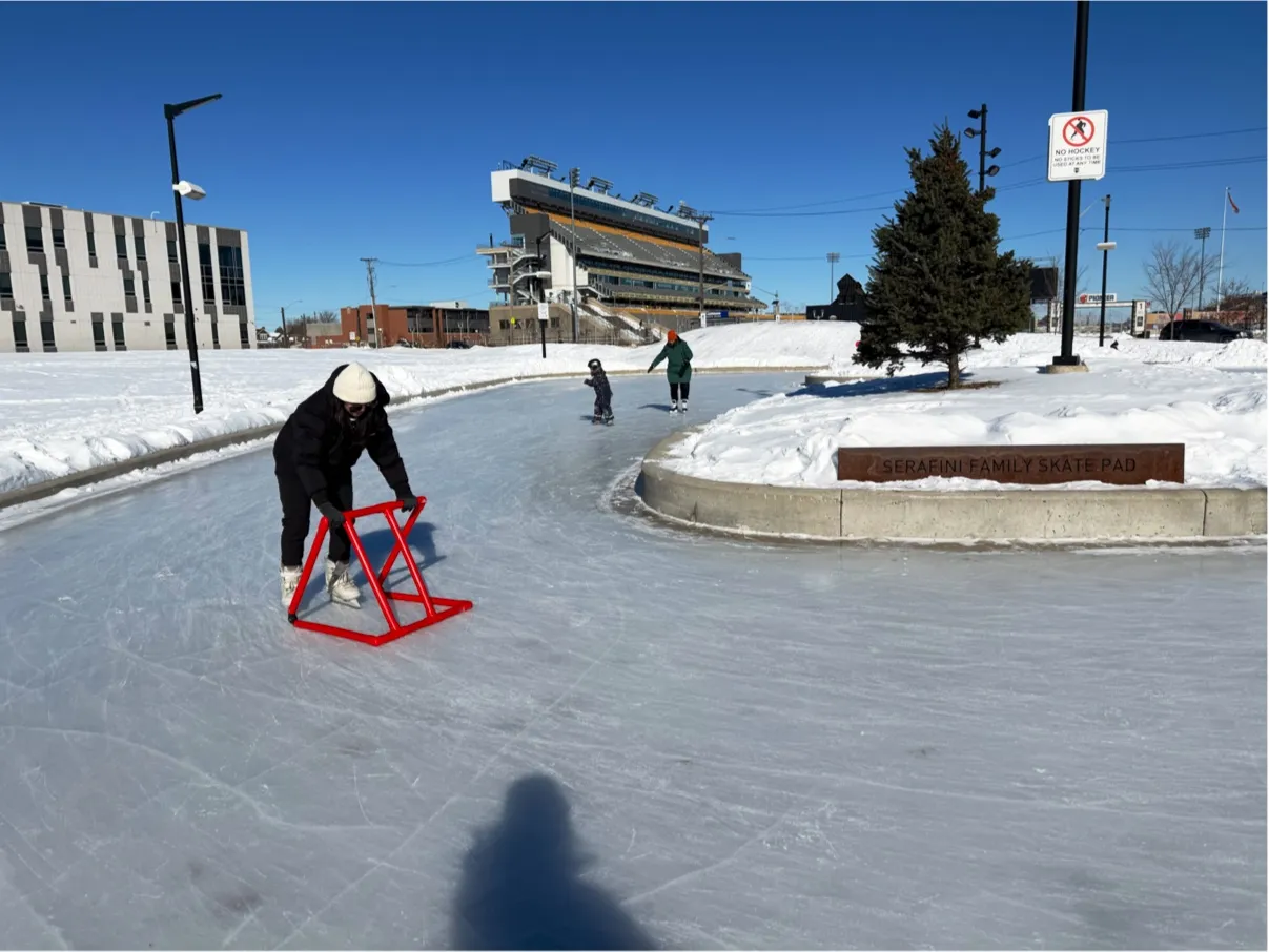 The curved ice pad at Bernie Morelli Recreation Centre in winter with a skater using a training aid and the stadium stands behind — Serafini Family Outdoor Ice Pad at Bernie Morelli Recreation Centre