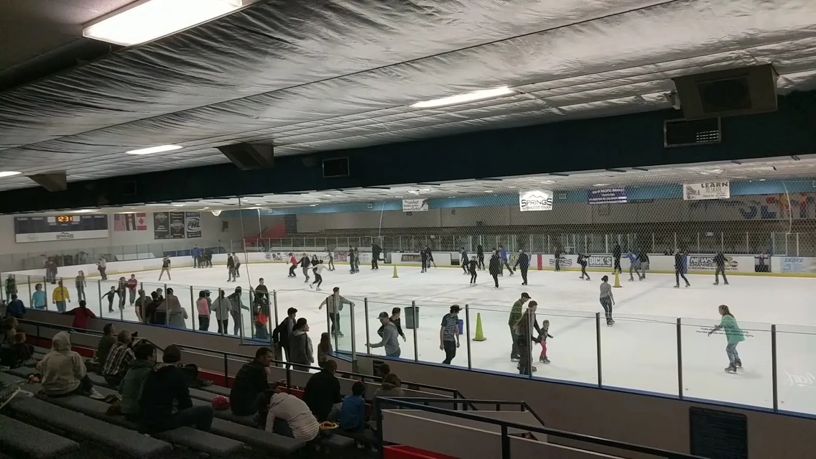 A public session seen from the bleachers across the glass, skaters spread across a wide white sheet under a long pale ceiling. — Sertich Ice Center