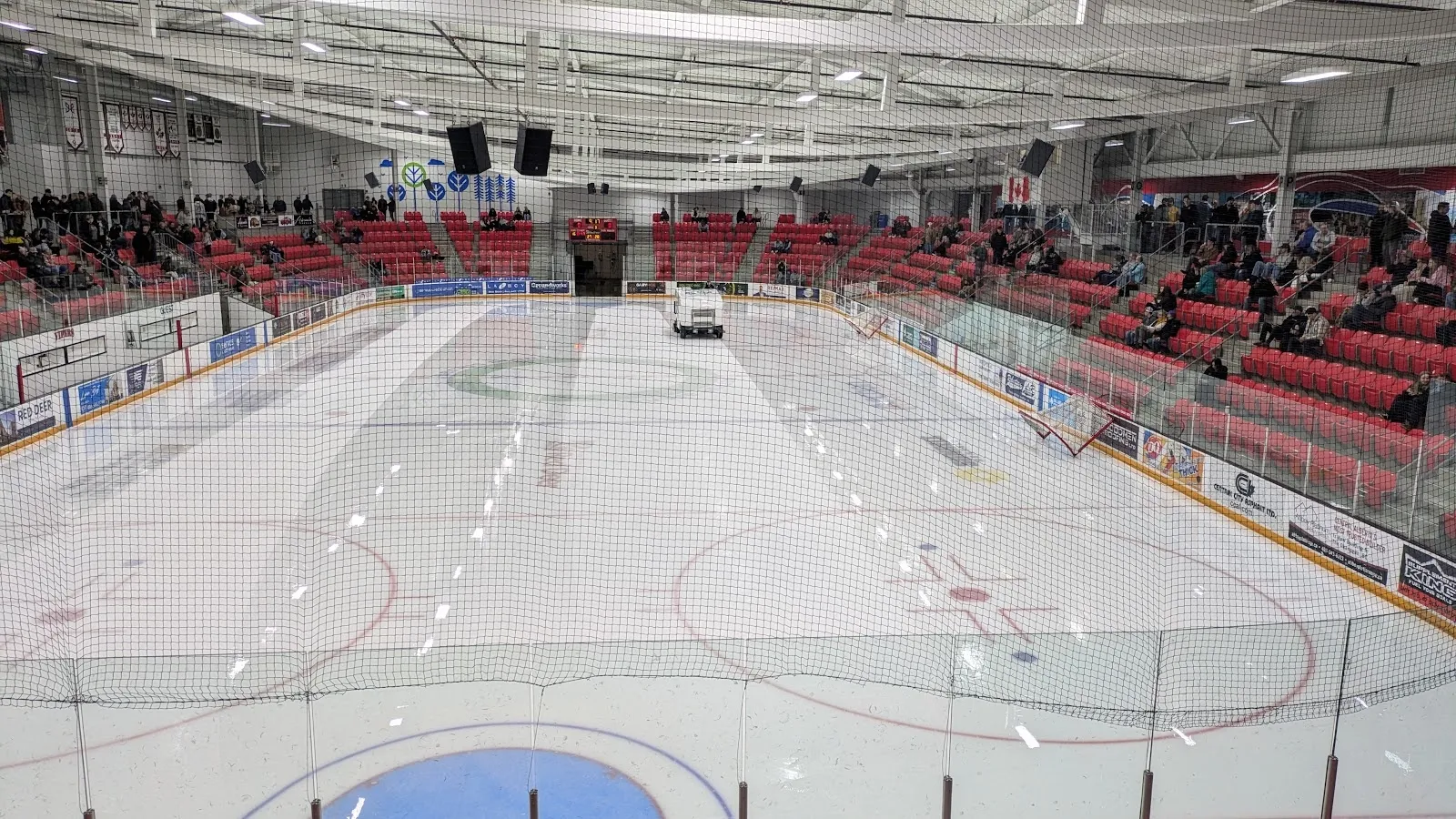 A wide view of the sheet with a resurfacer crossing the ice, red seating banked along the far side under a steel roof. — Servus Arena