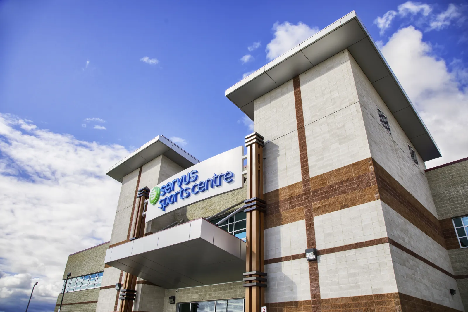 Las torres de entrada del Servus Sports Centre, de ladrillo y paneles claros, con el letrero verde de Servus Sports Centre sobre la marquesina, contra un cielo azul con nubes. — Servus Sports Centre