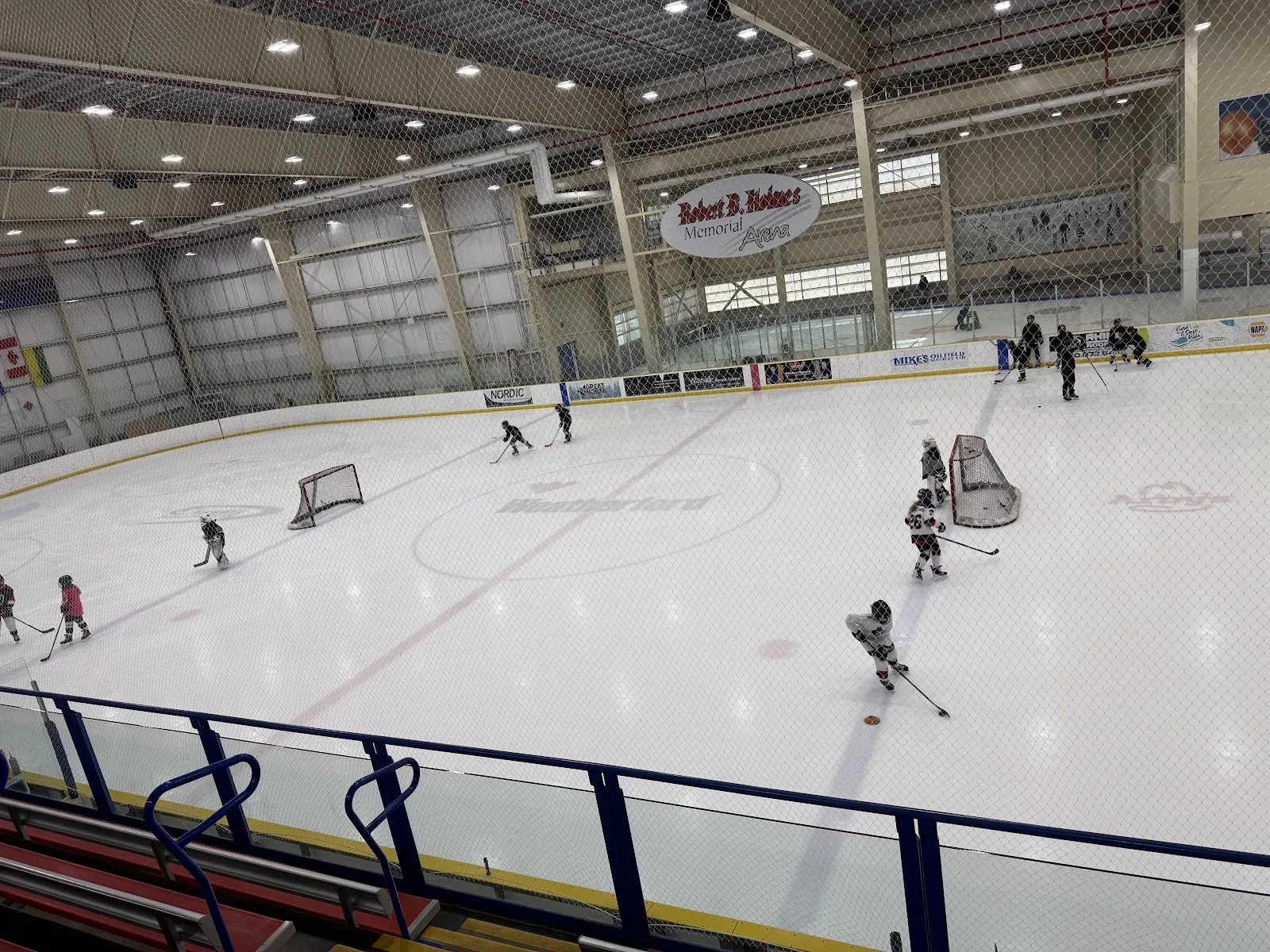 Un partido visto desde un barandal superior, con jugadores repartidos por una pista amplia con porterías en cada extremo bajo un alto techo con estructura de acero. — Servus Sports Centre