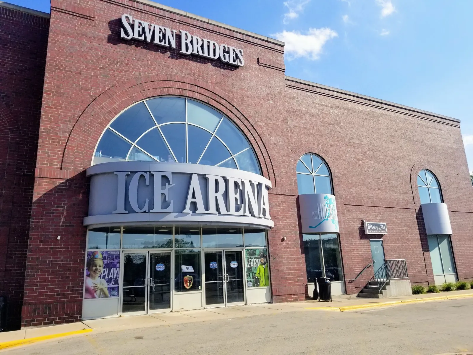 Storefront-style brick entrance with a large arched window, reading "Seven Bridges Ice Arena" above and "Ice Hockey" and "Whiskey Hill" signage beside the doors. — Seven Bridges Ice Arena
