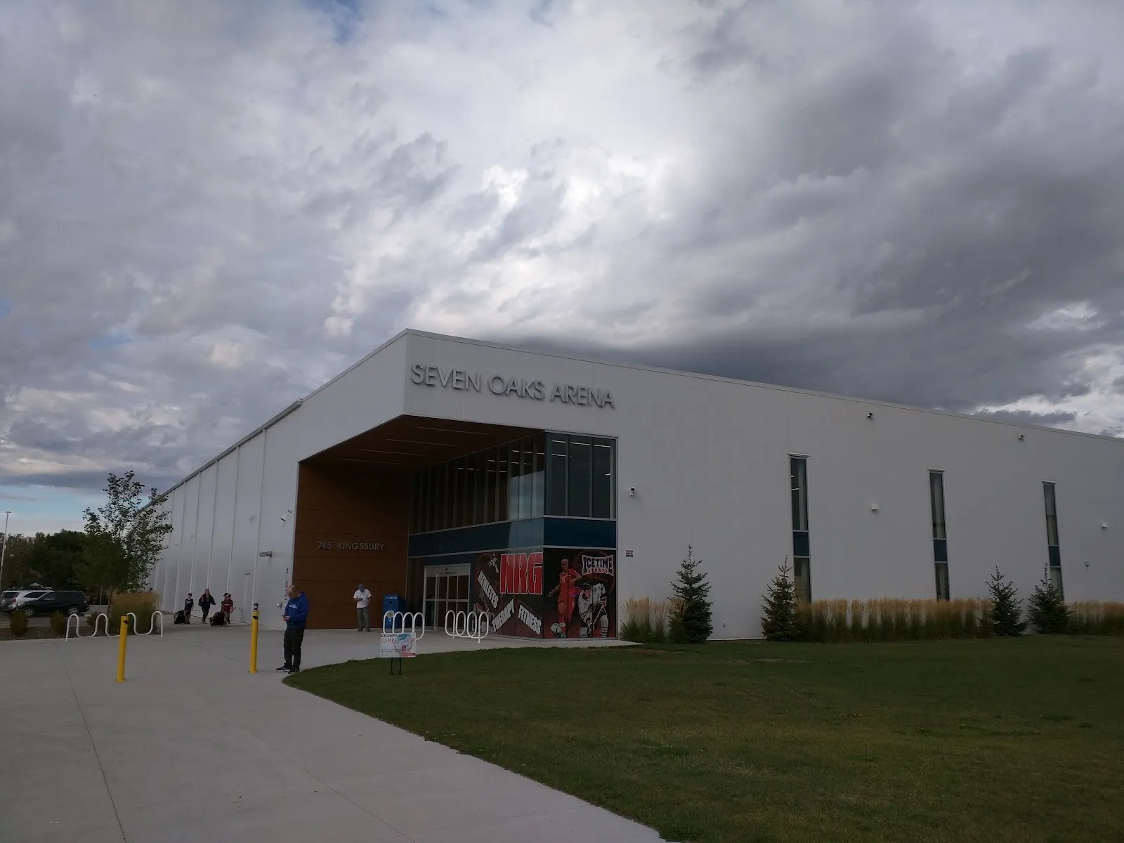 The white Seven Oaks Arena lettered above a recessed wood-lined entrance, tall grasses along the front and a few people near the doors. — Seven Oaks Arena