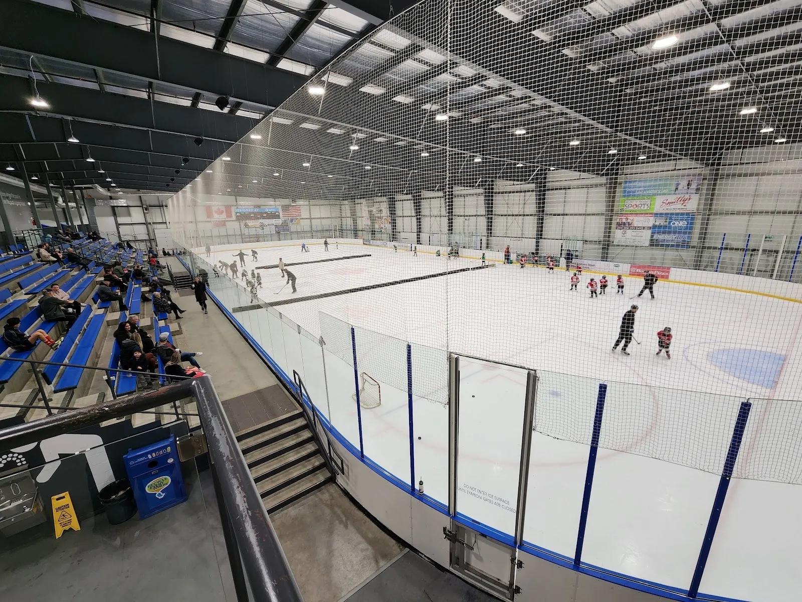 A wide view down the glass of the arena, players spread across a bright sheet with blue seating and a steel-trussed roof. — Seven Oaks Arena