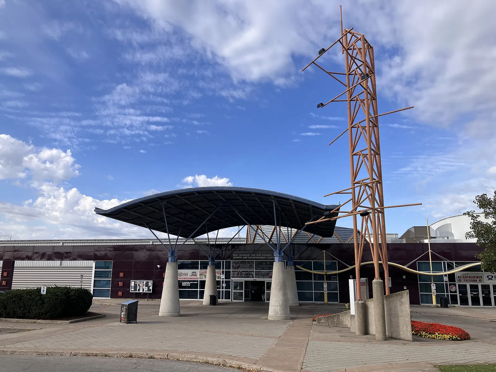 The glass-fronted entrance of the Seymour-Hannah Sports and Entertainment Centre in St. Catharines, under a curved canopy beside a tall wooden sculpture. — Seymour-Hannah Sports and Entertainment Centre