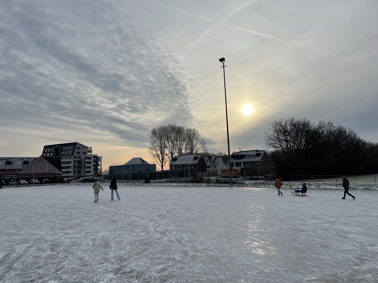 The flooded 's-Gravendeel rink under pale winter light, with a few skaters far off and houses along the edge. — IJsbaan 's-Gravendeel ('s-Gravendeelse IJsclub)