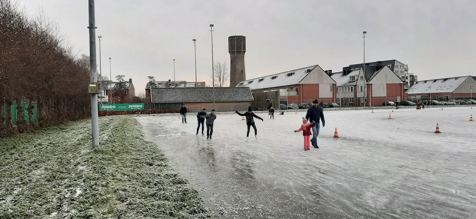 Skaters on the grey ice of the 's-Gravendeel rink with the club buildings and a water tower behind. — IJsbaan 's-Gravendeel ('s-Gravendeelse IJsclub)