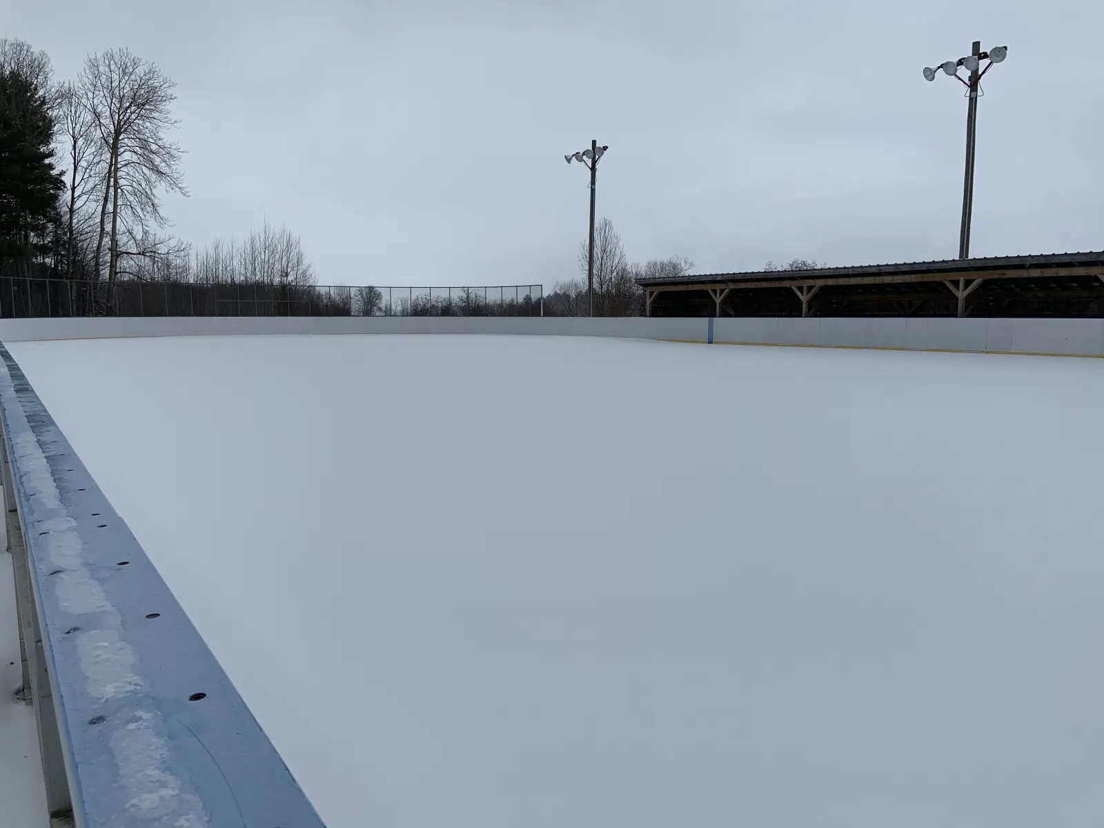 El hielo blanco y vacío de la pista exterior de Shady Nook a lo largo de sus vallas, con postes de focos y una gradería cubierta al fondo. — Shady Nook Outdoor Rink (Laurentian Valley)