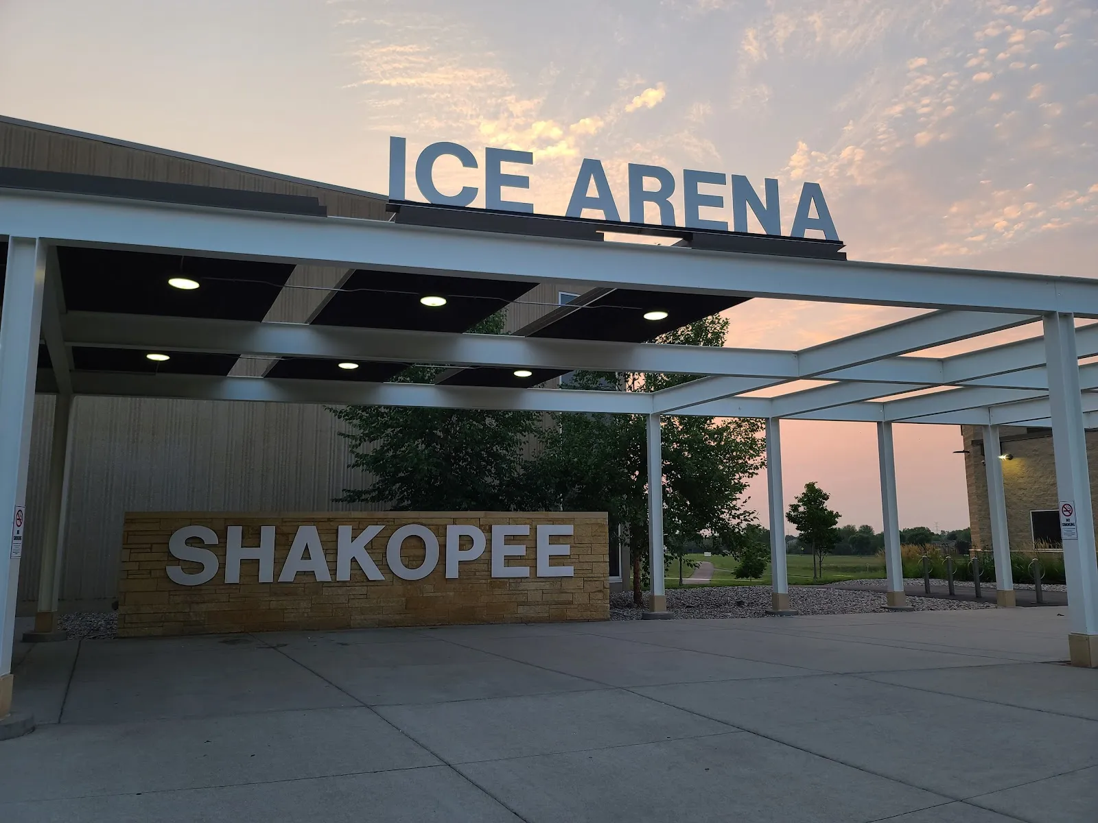 Entrance canopy at sunset with "ICE ARENA" lettering on the building fascia and a "SHAKOPEE" sign on the stone wall below it. — Shakopee Ice Arena