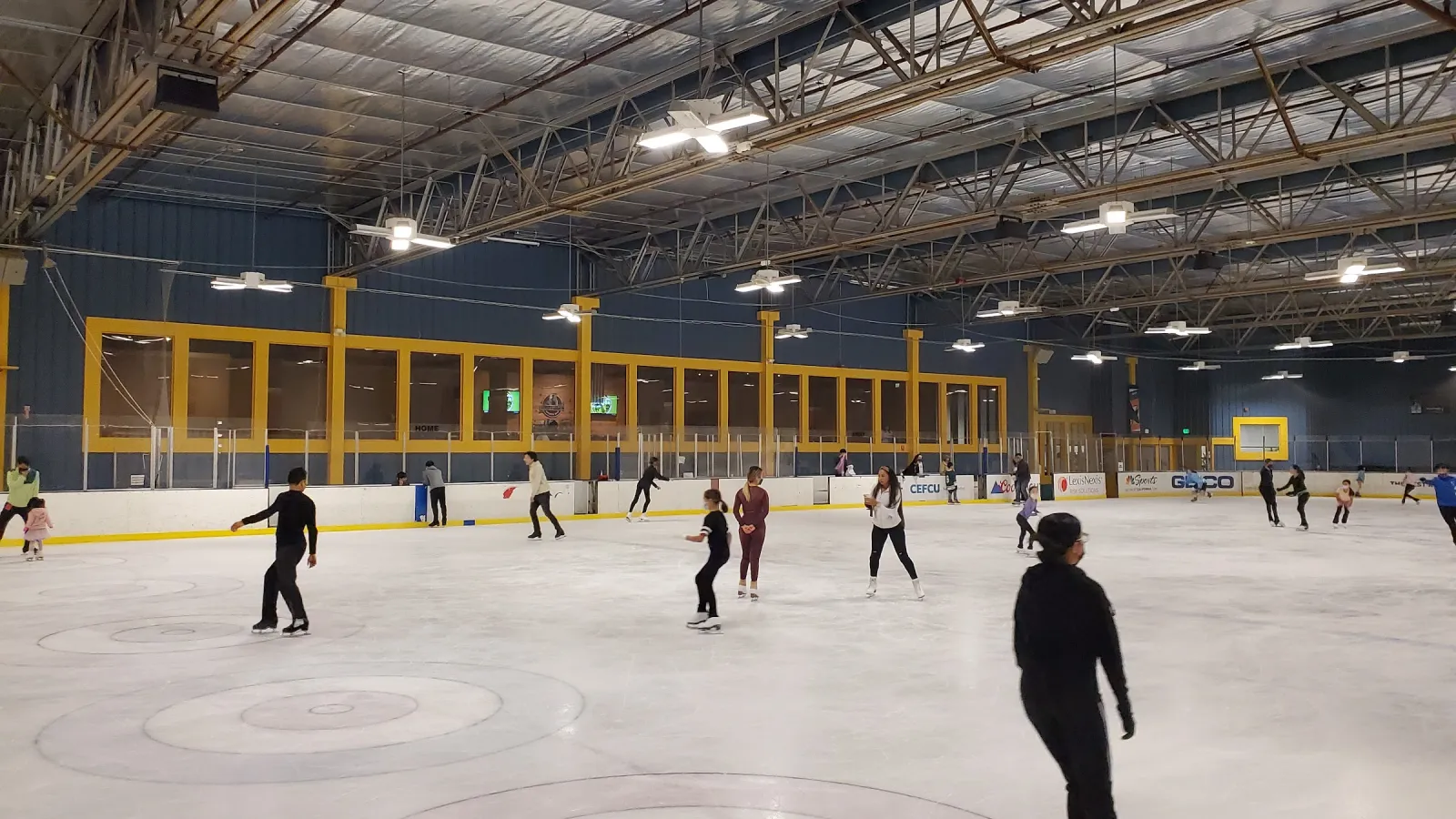 A public skate across a full sheet, seen from the ice, with the yellow-framed windows of the upper level and a grey steel roof. — Sharks Ice at Fremont