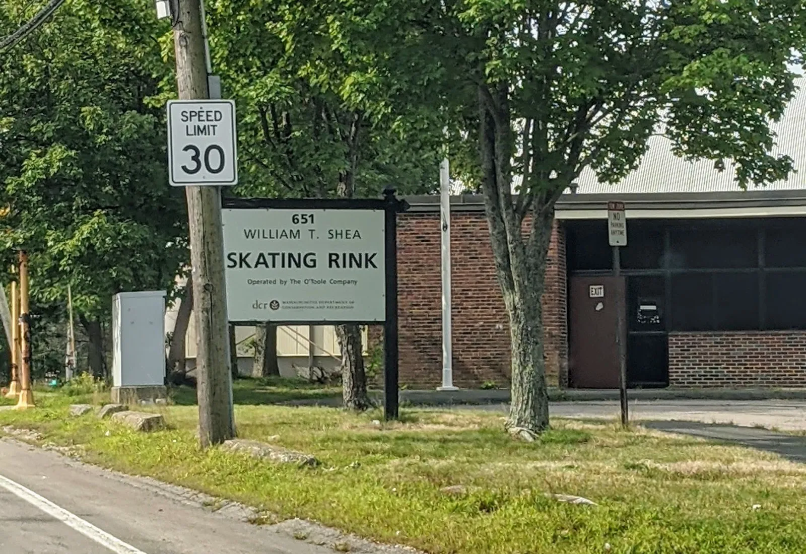 La William T. Shea Skating Rink desde Willard Street: el letrero 651 del DCR frente al bloque de entrada de ladrillo, con el techo curvo de la pista detrás de los árboles — Shea Memorial Rink