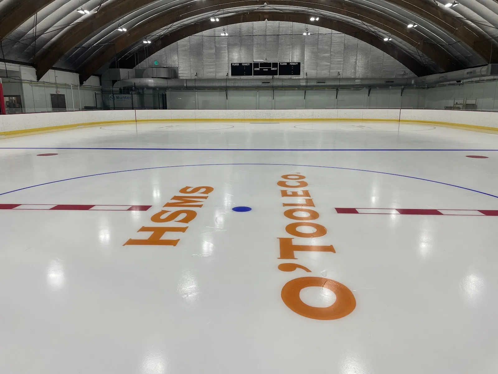 Toda la pista vacía bajo un techo en arco de madera y metal, con un marcador, vallas con remate amarillo y logotipos de negocios locales pintados en el centro de la pista. — Shea Memorial Rink