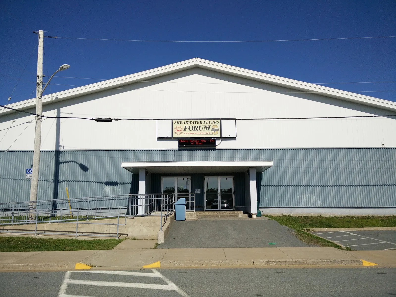The front of the Shearwater Flyers Forum, a white-gabled arena with blue-grey cladding, a small canopy over its glass doors and a wheelchair ramp, seen from across the street under a deep blue sky. — Shearwater Arena