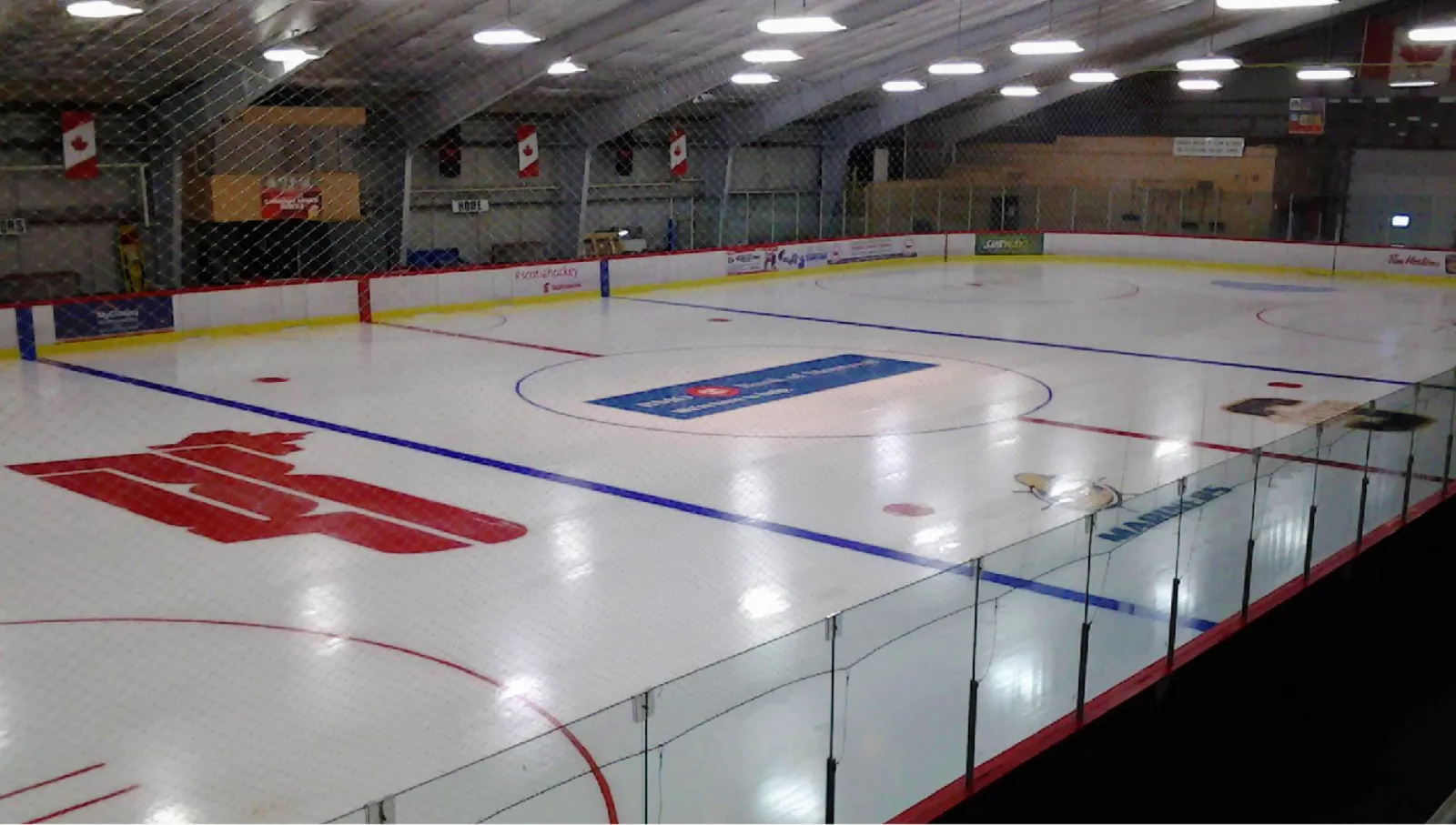 Full ice sheet inside Shearwater Arena, viewed from the glass at one end; overhead lighting, red Air Force roundel painted at centre ice, Canadian flags on the far wall, dasher-board ads for Scotiabank, Tim Hortons and Subway. — Shearwater Arena