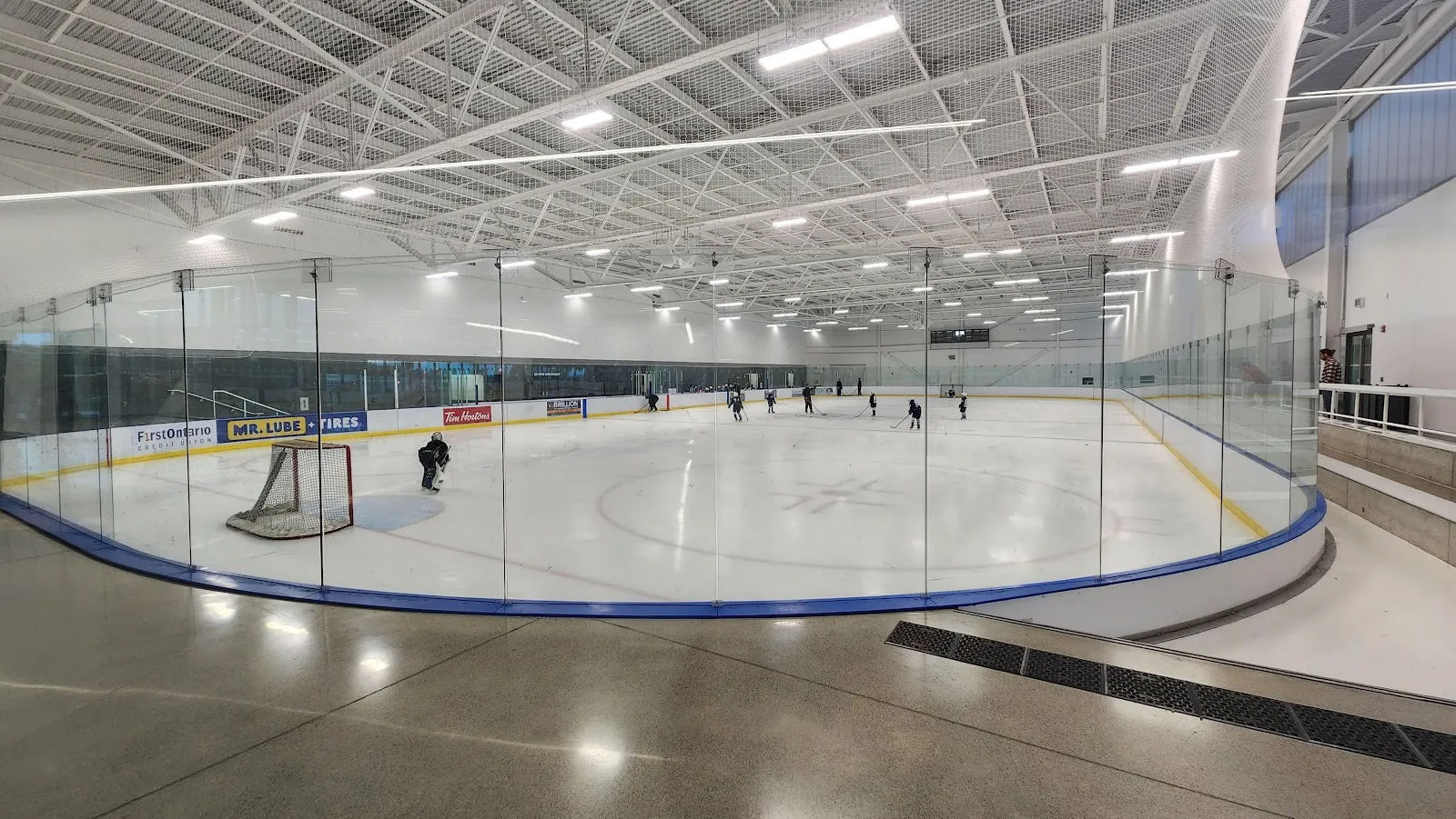 One of the two rinks seen through the glass from the lobby side, with skaters on the ice and a curved blue board line. — Sherwood Community Centre