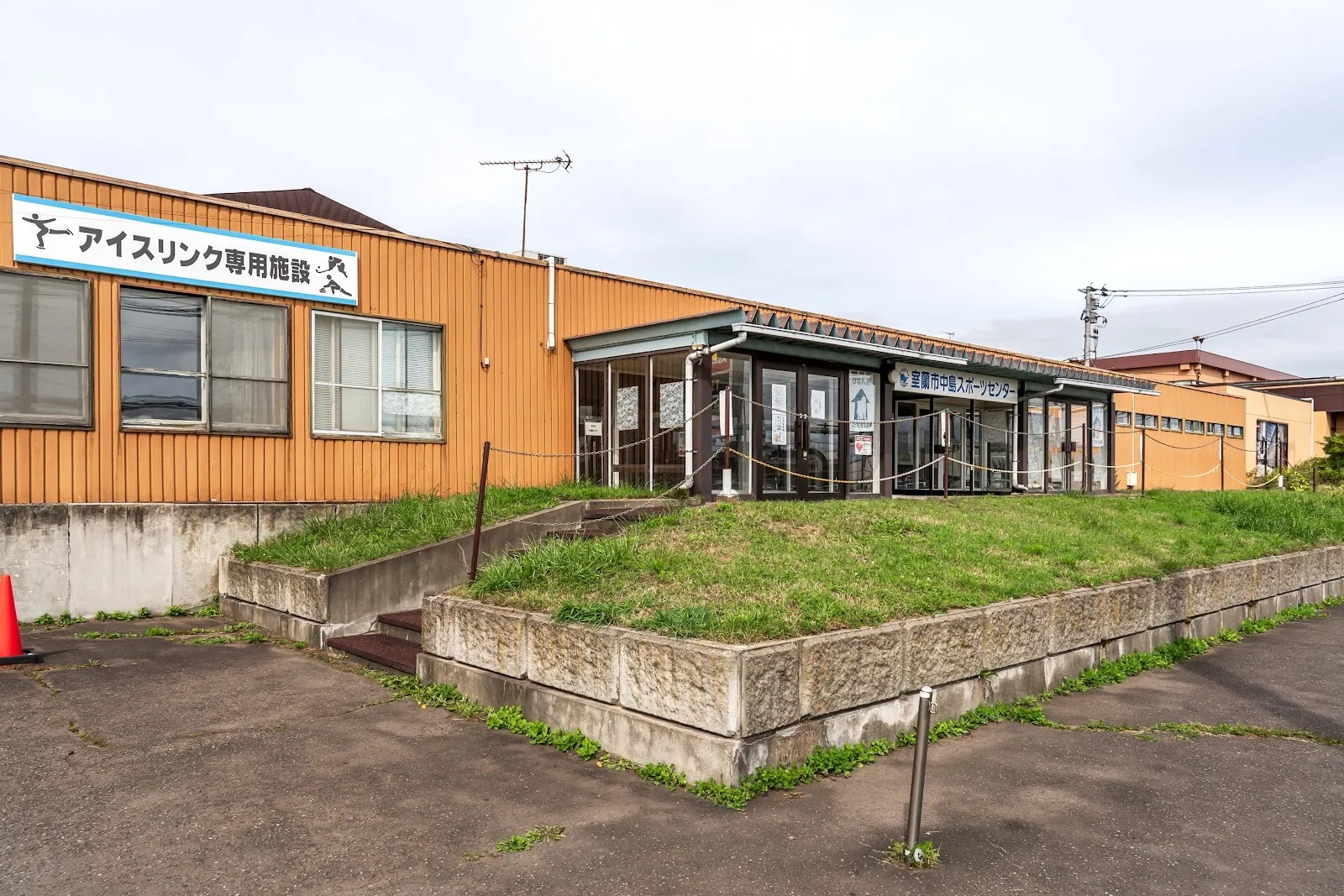 The orange-clad entrance building of the Muroran ice rink, with its blue ice-rink-only facility sign, a grassy bank and a concrete retaining wall in front. — Shinwa Sangyo Ice Arena Muroran