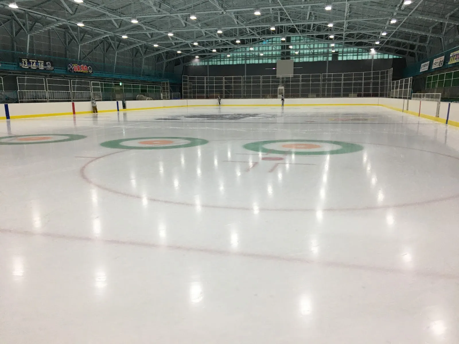 The empty ice sheet of Shinwa Sangyo Ice Arena Muroran with green face-off circles and red lines marked on the white ice, under a steel roof with a green band of windows. — Shinwa Sangyo Ice Arena Muroran