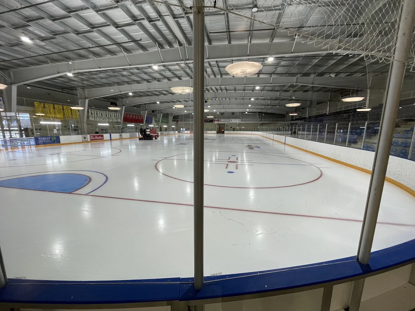 El hielo vacío de tamaño NHL desde la banca, con la pulidora de hielo en el extremo opuesto y mantas bajo el techo. — Shores Recreation Centre (Forest)