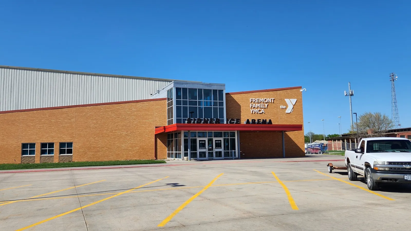 The brick front of Sidner Ice Arena at the Fremont Family YMCA, with the red canopy over the entrance, the arena name above the doors and a large parking lot in front. — Sidner Ice Arena