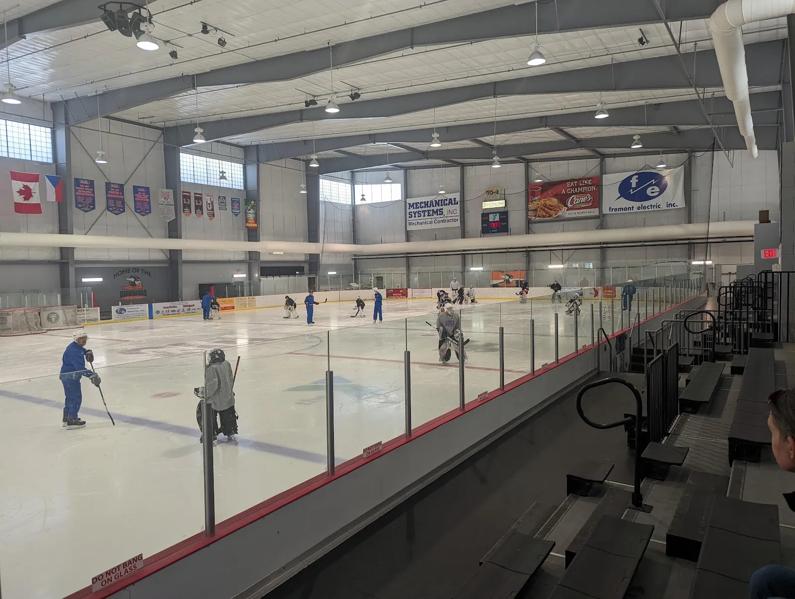 A wide view down the glass of the sheet, a few skaters far across the ice and banners along the upper wall under a steel roof with a spectator walkway on the right. — Sidner Ice Arena