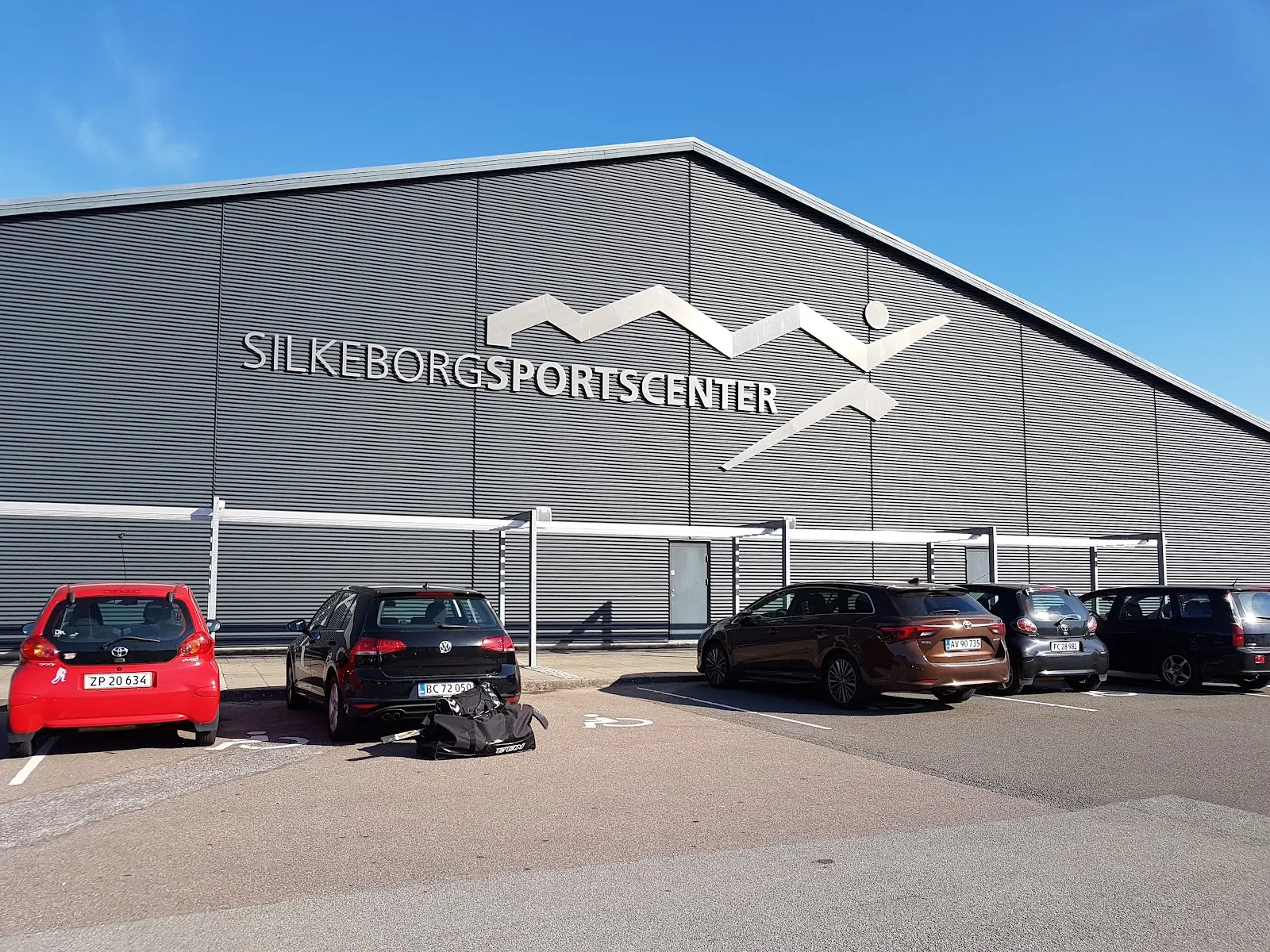 The grey front of Silkeborg Sportscenter, the sports centre that holds the skating hall, with its name and logo on the wall above a row of parked cars under a blue sky. — Silkeborg Skøjtehal