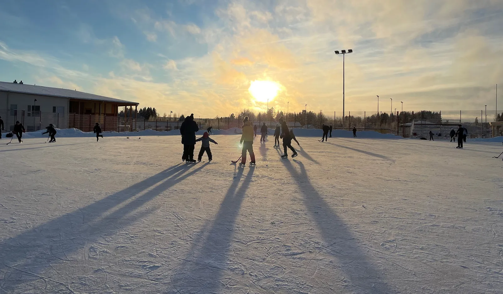 Pelaajia ja luistelijoita Siltatien tekojäärata Ylöjärvellä auringonlaskussa, jäällä pitkät varjot ja taustalla liikuntarakennus. – Siltatien tekojää (Ylöjärvi)