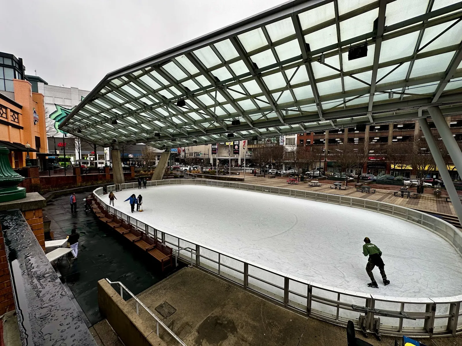 The covered outdoor ice at the Silver Spring Ice Skating Pavilion with a lone skater under the glass-and-steel roof and the plaza buildings behind. — Silver Spring Ice Skating at Veterans Plaza