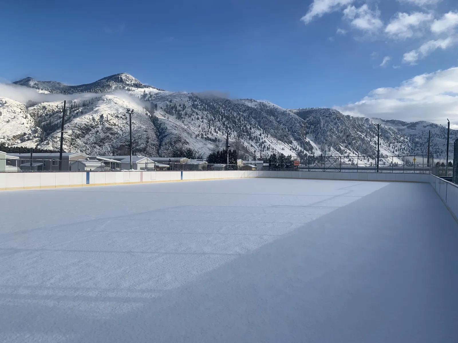 La pista exterior refrigerada del Similkameen Recreation Centre, en Keremeos, en un luminoso día de invierno, con hielo limpio dentro de las vallas y montañas cubiertas de nieve al fondo. — Similkameen Recreation Centre Outdoor Rink